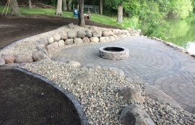 Circular paver patio around a raised fire pit with a boulder retaining wall running along the backside