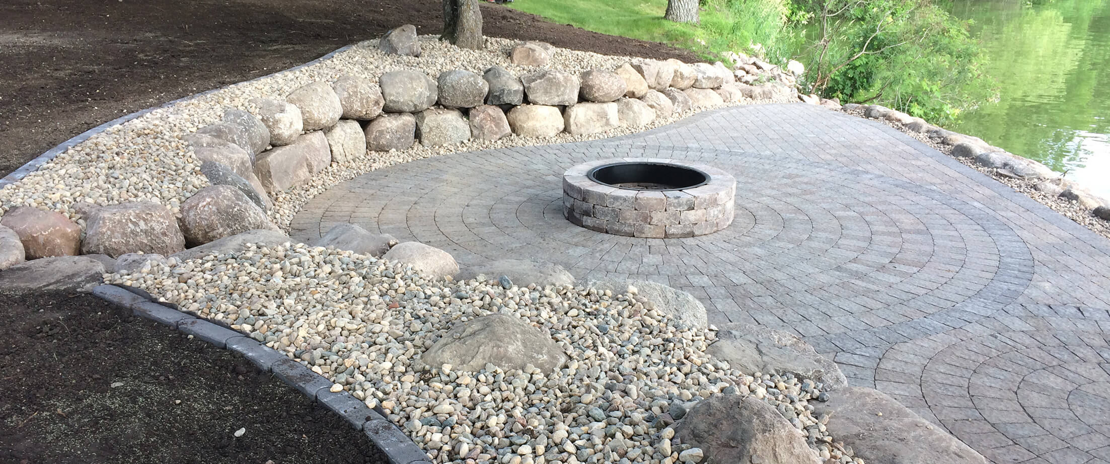 Circular paver patio around a raised fire pit with a fieldstone boulder retaining wall around the perimeter